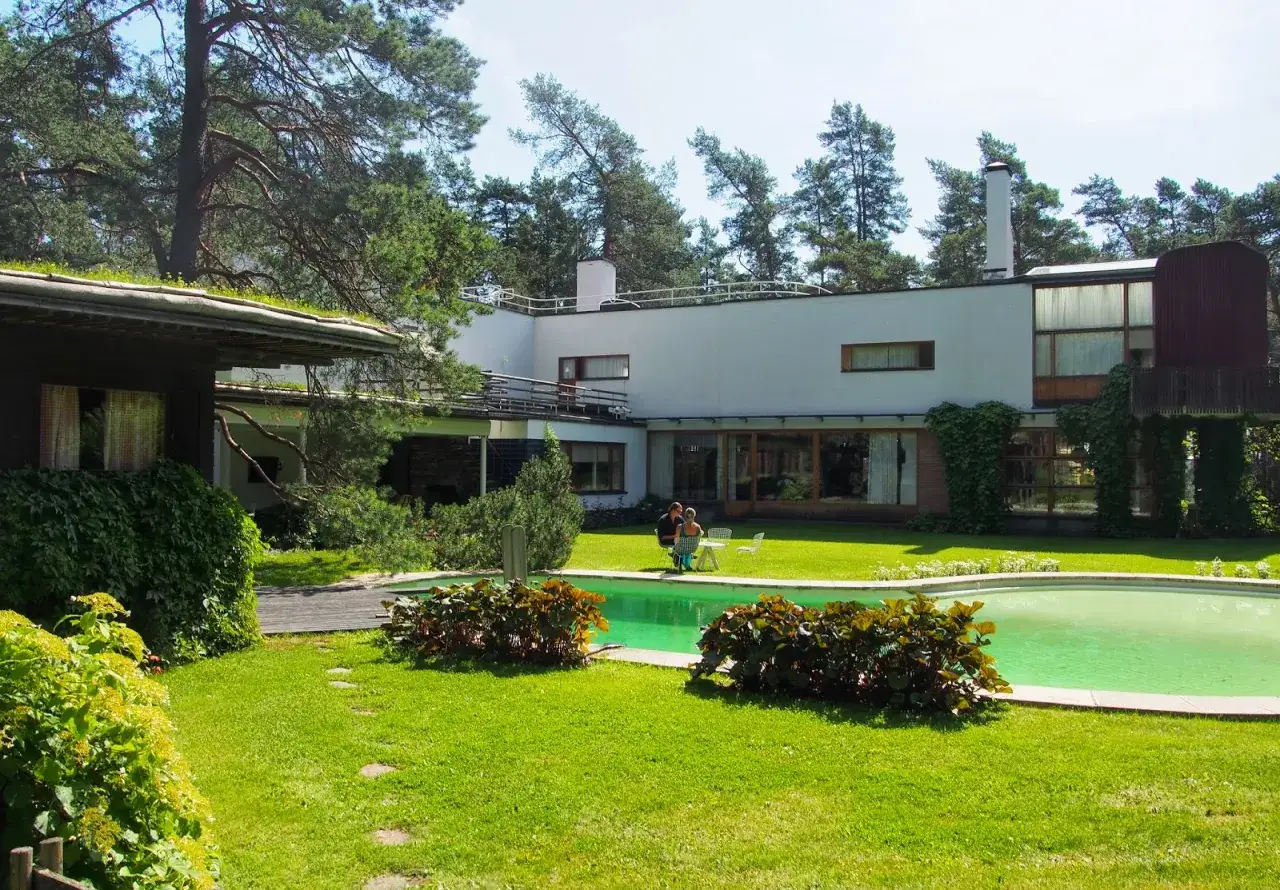 Villa Mairea, z basenem i ogrodem, otoczona sosnami. Dwie osoby siedzą przy stoliku na trawniku.