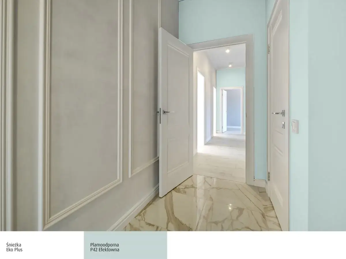 Jasny korytarz z białymi drzwiami i eleganckimi sztukateriami. Płynne przejście do kolejnych pomieszczeń sugeruje, jaka najlepsza farba do przedpokoju – jasna i odporna.