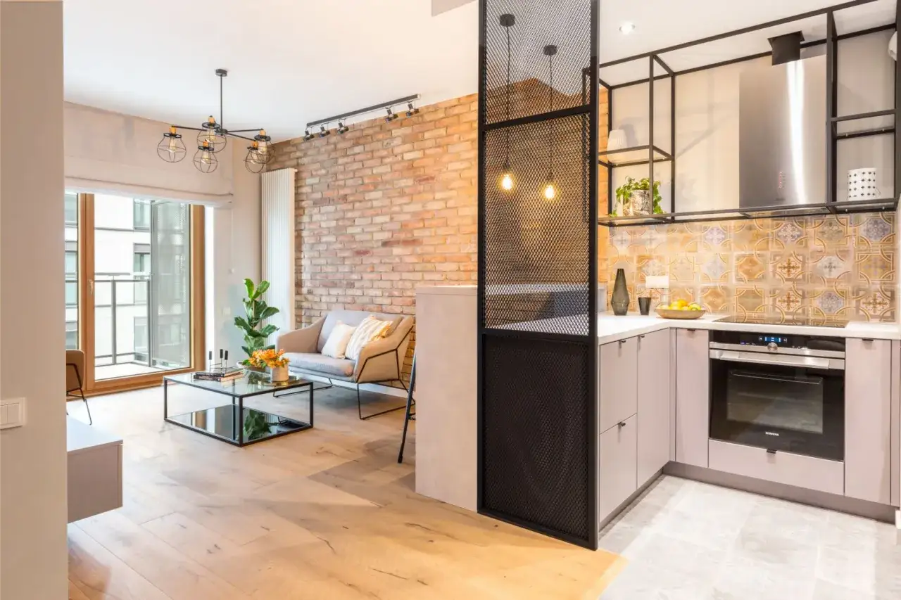 Nowoczesny **styl loftowy w bloku**: ceglana ściana, szara kanapa, szklany stolik i kuchnia z metalowymi elementami.