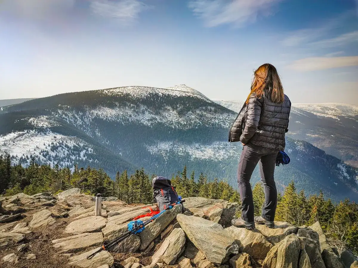 Kobieta na skale podziwia zimowy krajobraz. Na pierwszym planie plecak i kijki trekkingowe. W oddali widać ośnieżone g&oacute;ry i przełęcz okraj.