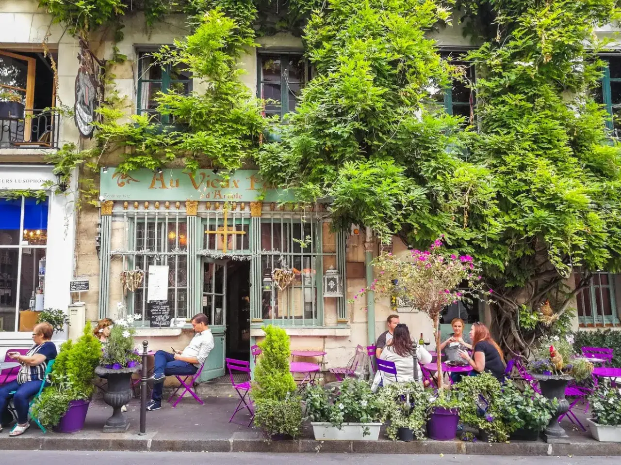 Urocza kawiarnia w 14 dzielnicy Paryża, otoczona zielenią, z ludźmi siedzącymi przy fioletowych stolikach.