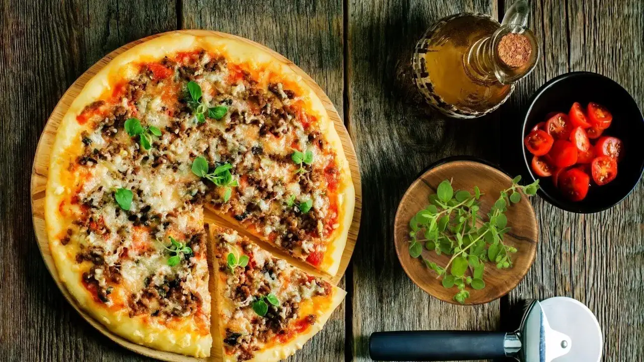 Pyszna pizza z mięsem mielonym, posypana świeżym oregano. Obok miseczka pomidork&oacute;w i buteleczka oliwy. Idealna na wiecz&oacute;r z przyjaci&oacute;łmi.