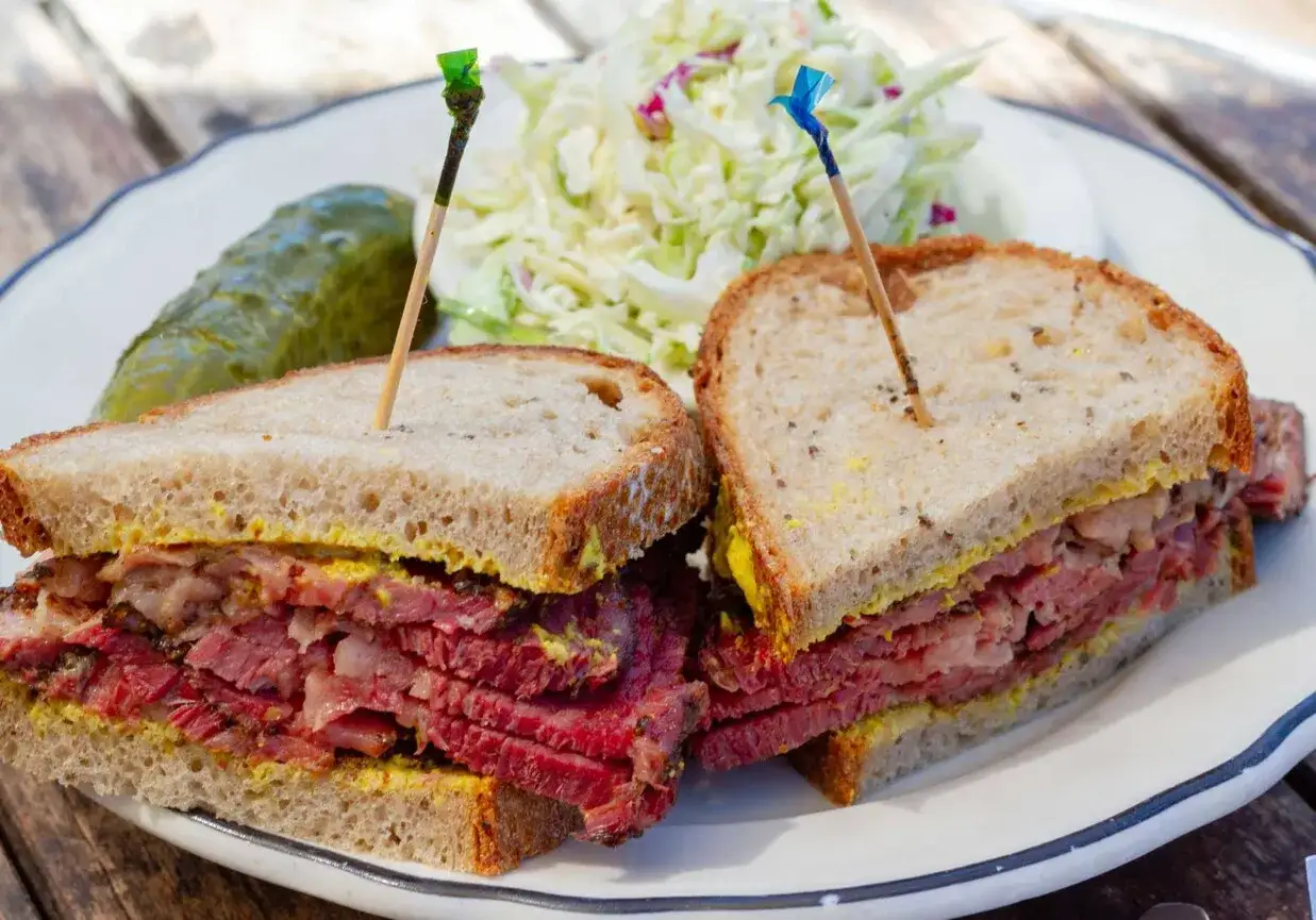 Pyszna kanapka z pastrami, obficie przełożona mięsem, z musztardą, obok sur&oacute;wka i og&oacute;rek kiszony.