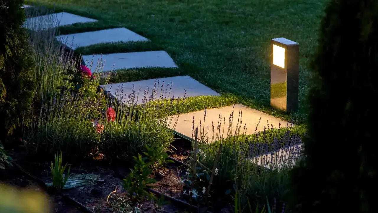 Ścieżka z płyt kamiennych w ogrodzie, oświetlona lampami. Widać lawendę i r&oacute;że. Lampy ogrodowe rozmieszczone co kilka metr&oacute;w, tworzą magiczny nastr&oacute;j.