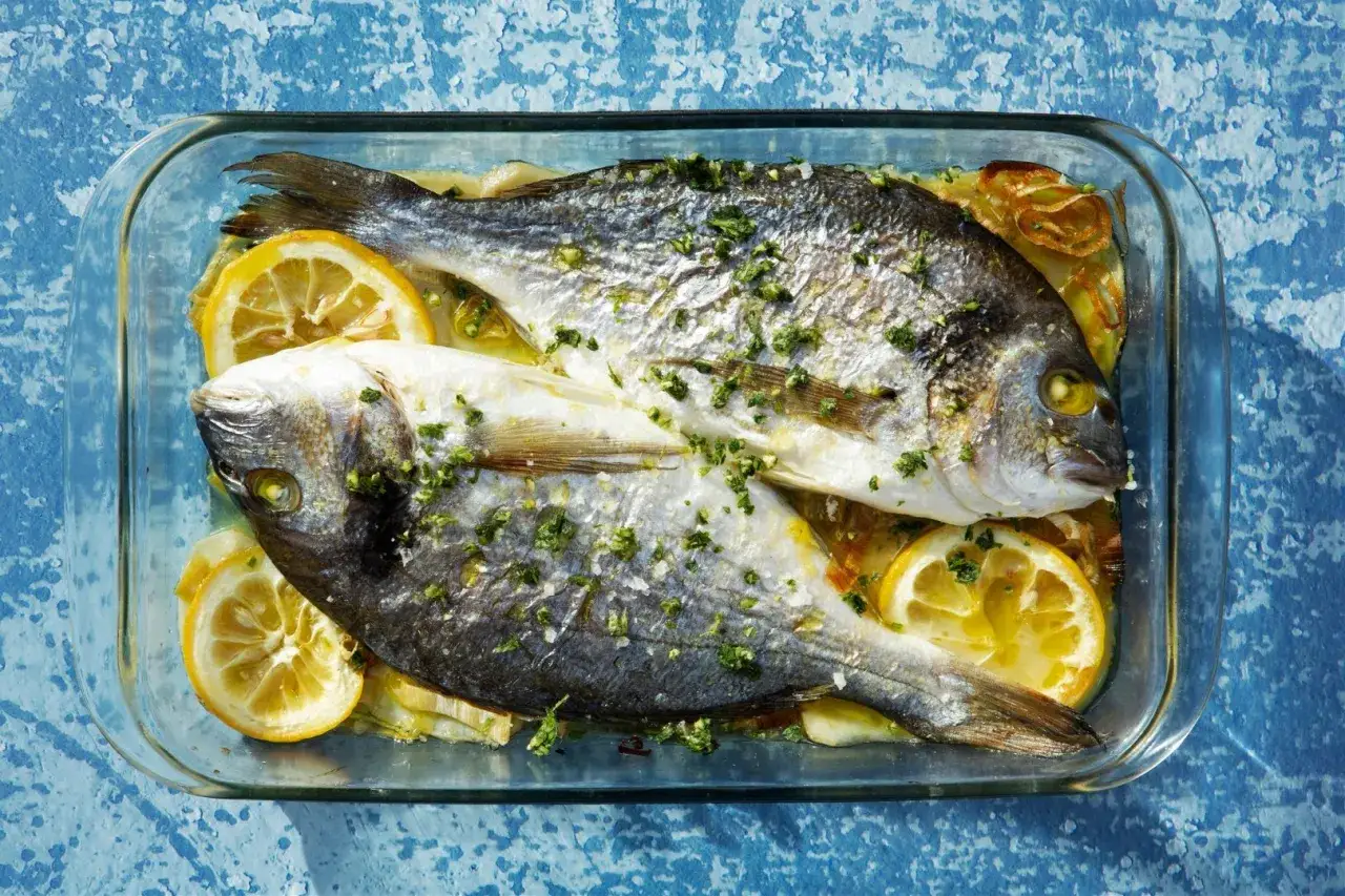 Dwie dorady z piekarnika, skropione ziołami i cytryną, w szklanym naczyniu.