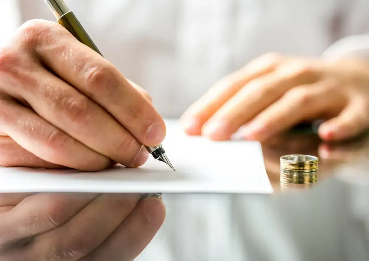 Dłoń pisze piórem na kartce, obok leży obrączka. To symboliczny moment, gdy ktoś składa pozew o rozwód.