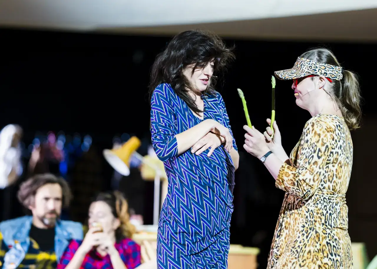 Dwie kobiety na scenie w Warszawie, jedna w niebieskiej sukience, druga w panterce, trzymają szparagi. Spektakl 