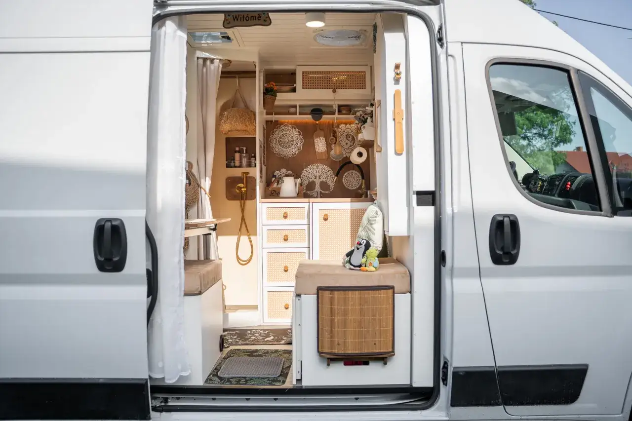 Przytulne wnętrze białego busa kampera z rattanowymi frontami szafek, ozdobami na ścianie i pluszowymi zabawkami.