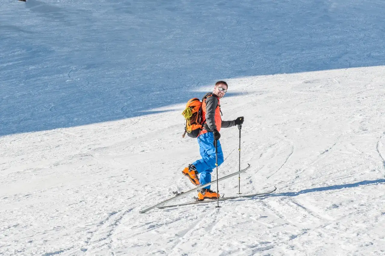 Uśmiechnięty mężczyzna w pomarańczowej kamizelce i niebieskich spodniach na nartach, gotowy do ski tury. Plecak i kijki wskazują na g&oacute;rską przygodę.