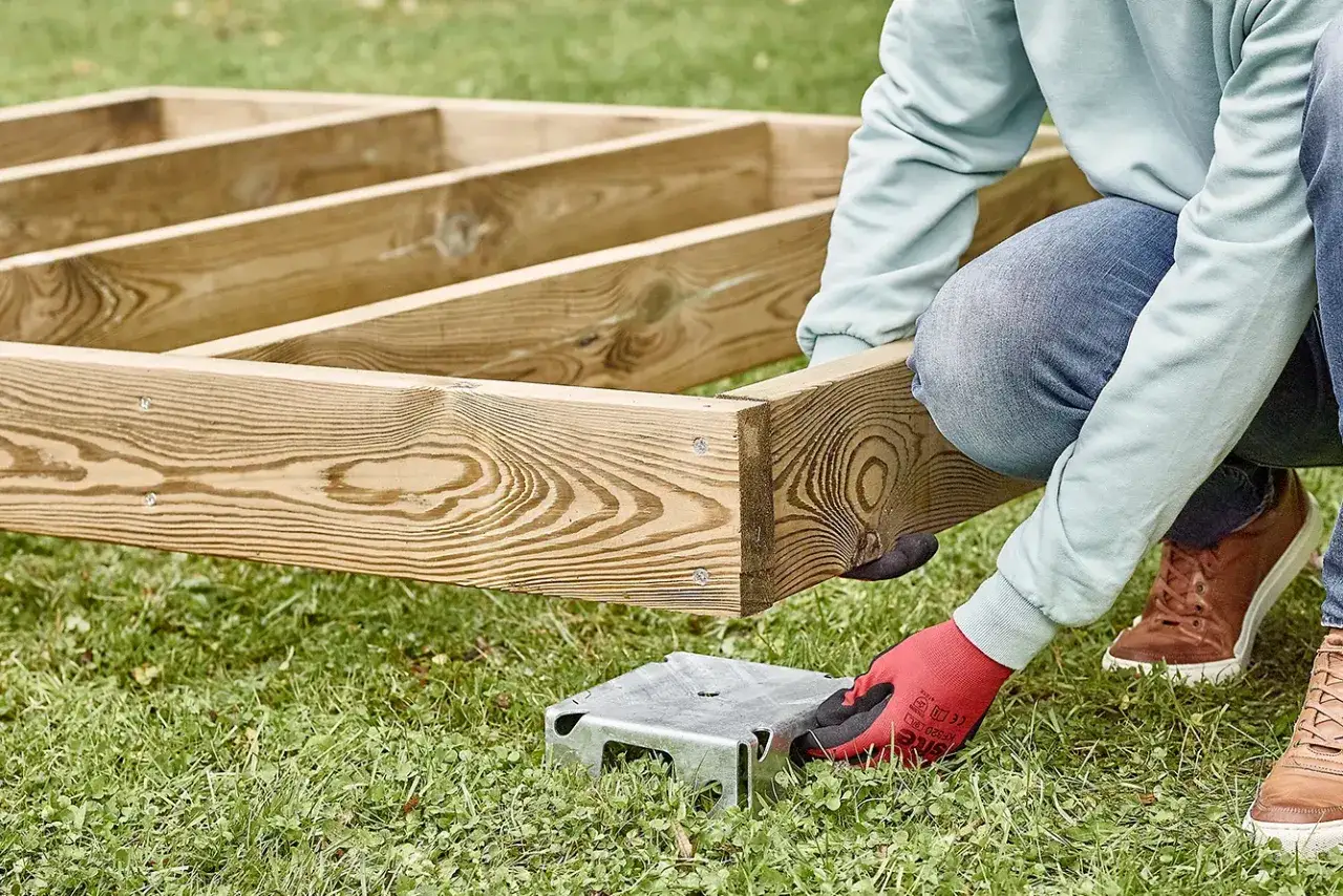 Budowanie podłoża pod domek narzędziowy. Drewniana konstrukcja opiera się na metalowych wspornikach na trawie.