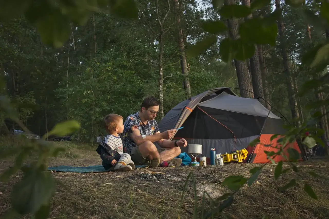 Ojciec z synkiem siedzą przy namiocie, planując, gdzie pod namiot rozbić obóz. Wokół las i sprzęt kempingowy.