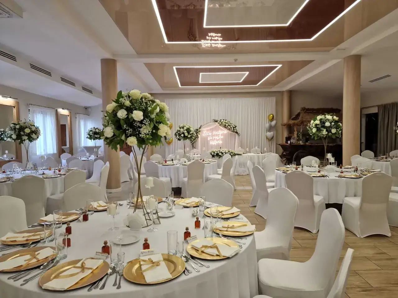 Elegancka sala weselna na rancho pod bocianem, udekorowana białymi kwiatami i złotymi akcentami. Stoły nakryte białymi obrusami, ozdobione złotymi talerzami i świecami.