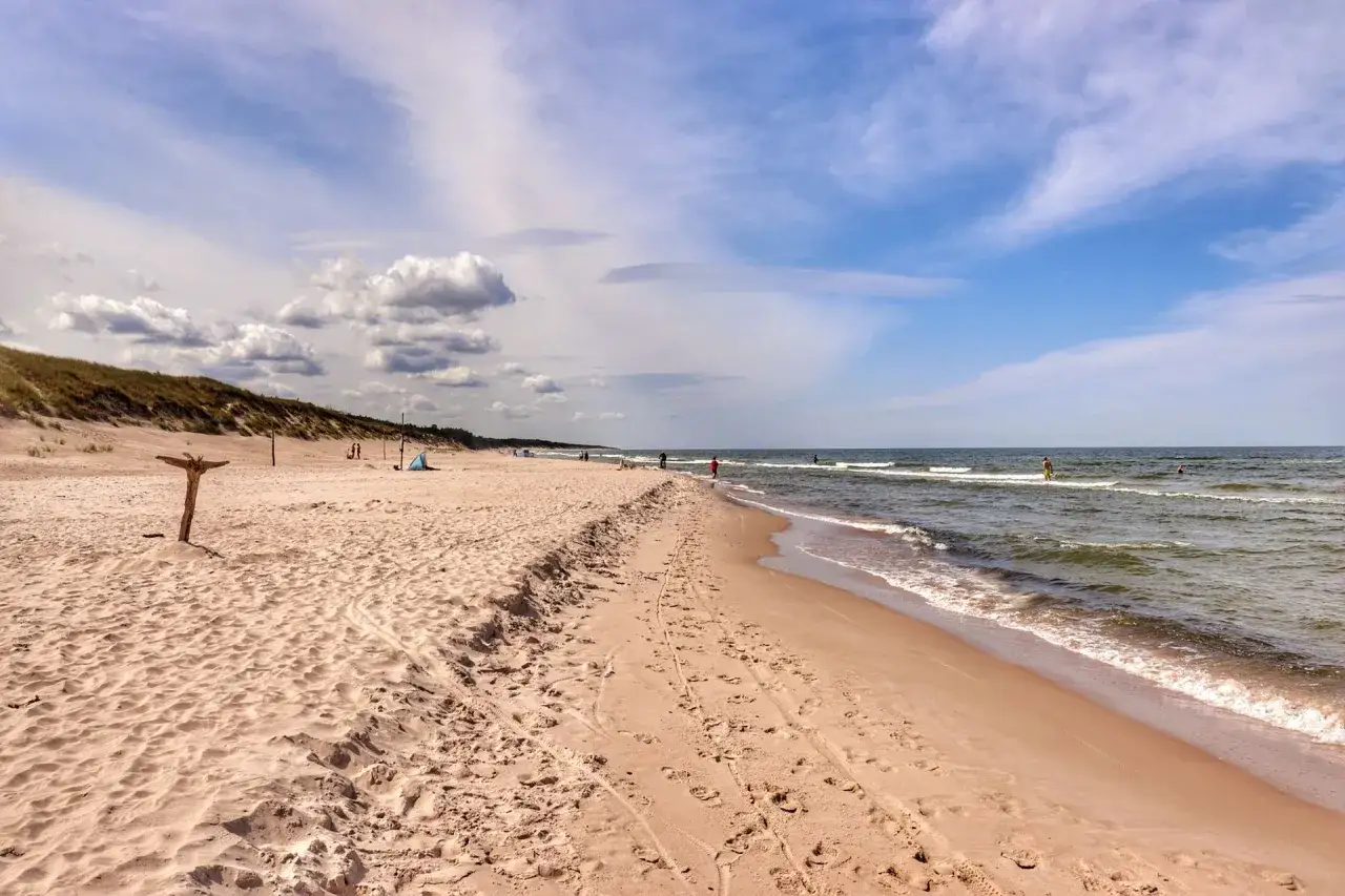 Poddąbie nad morzem: piaszczysta plaża, wydmy porośnięte trawą, spokojne morze i błękitne niebo z chmurami. Kilka osób spaceruje brzegiem.