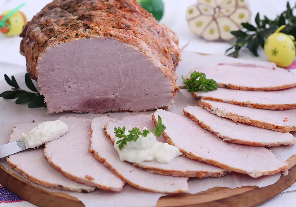 Soczysta szynka pieczona w rękawie, pokrojona w plastry, z chrzanem i natką pietruszki. Idealna na wielkanocny st&oacute;ł.