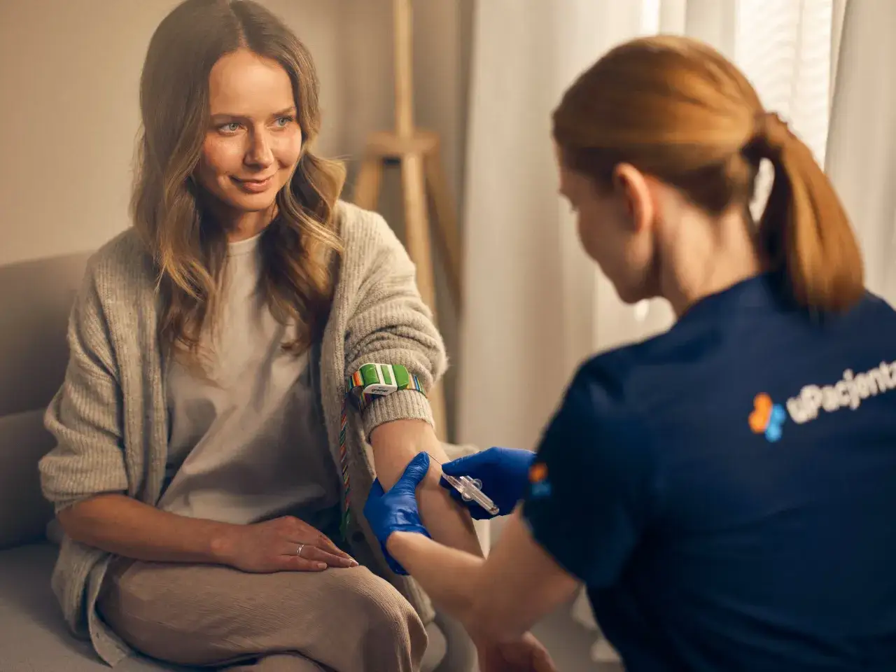 Uspokojona pacjentka uśmiecha się podczas pobierania krwi. Pielęgniarka w niebieskich rękawiczkach wykonuje zabieg, pokazując jak nie zemdleć przy pobieraniu krwi.