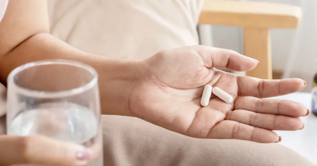 Dłoń trzyma dwie białe kapsułki, zastanawiając się, czy probiotyk przed czy po antybiotyku jest lepszy. Obok szklanka wody.