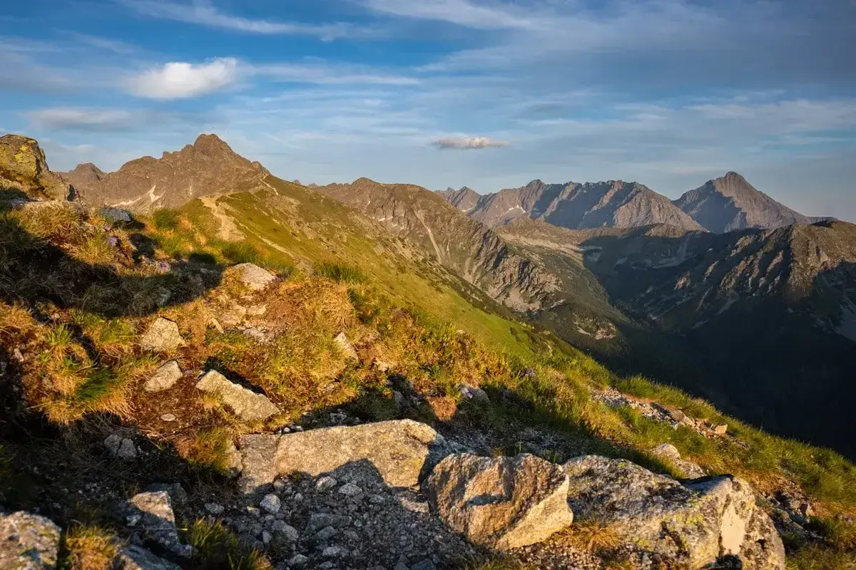 Widok z Kasprowego Wierchu w Zakopanem. Słoneczny dzień, g&oacute;ry w oddali, ścieżka wśr&oacute;d traw i kamieni.