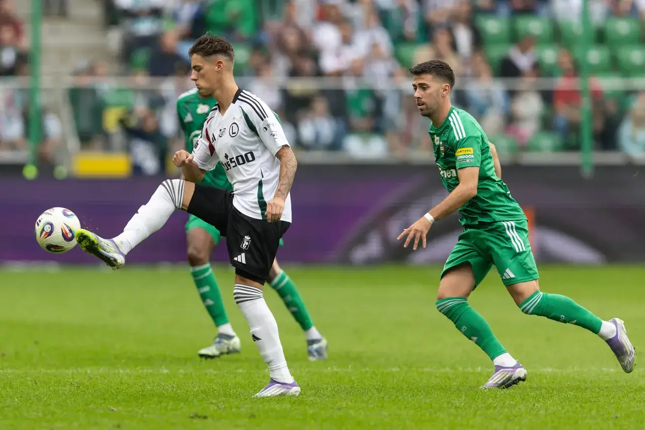 Piłkarz Legii Warszawa w białej koszulce kopie piłkę, podczas gdy zawodnik Pogoni Szczecin w zielonym stroju obserwuje grę.