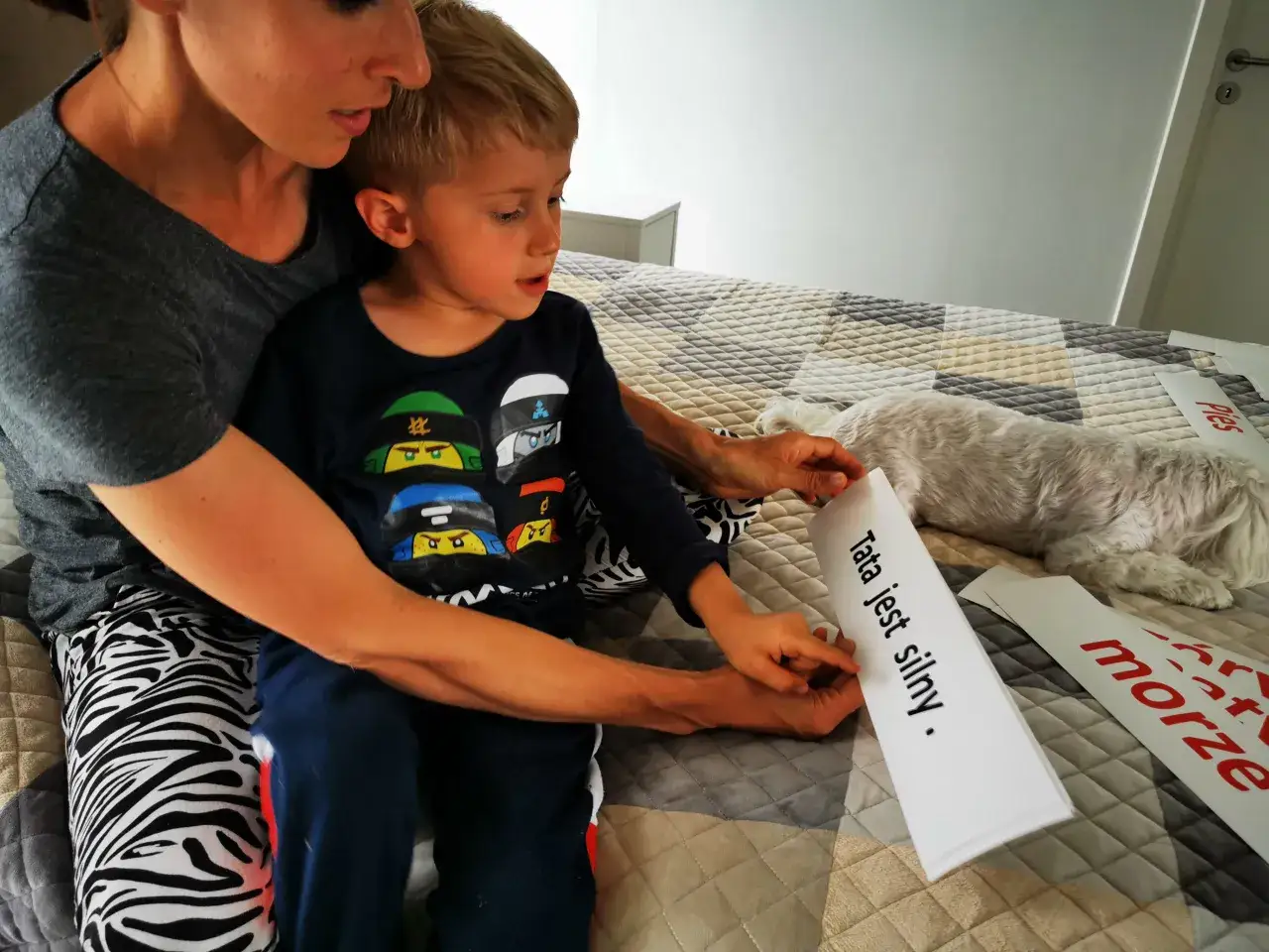 Mama pomaga 7 latkowi w nauce czytania. Chłopiec wskazuje palcem na kartkę z napisem 