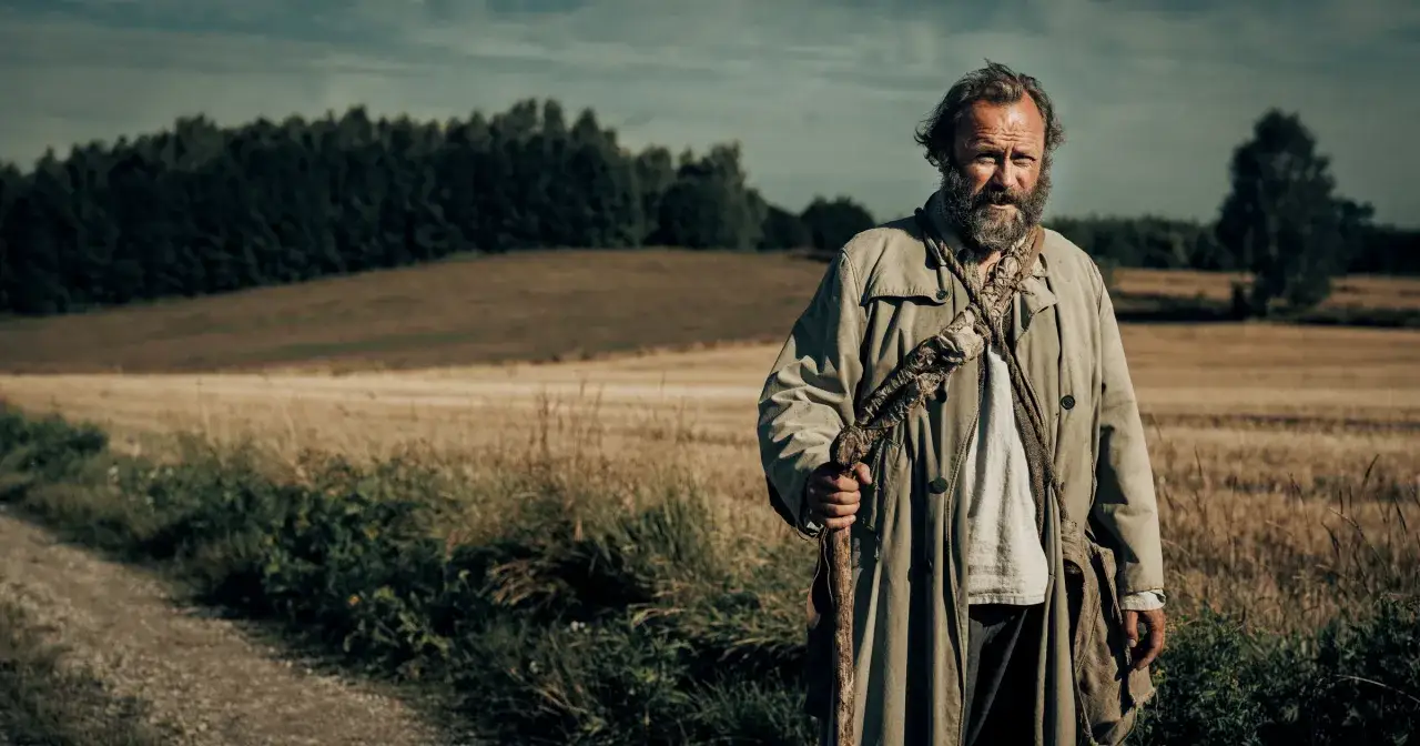 Stary znachor z laską i torbą na ramieniu, w płaszczu, stoi na polnej drodze.