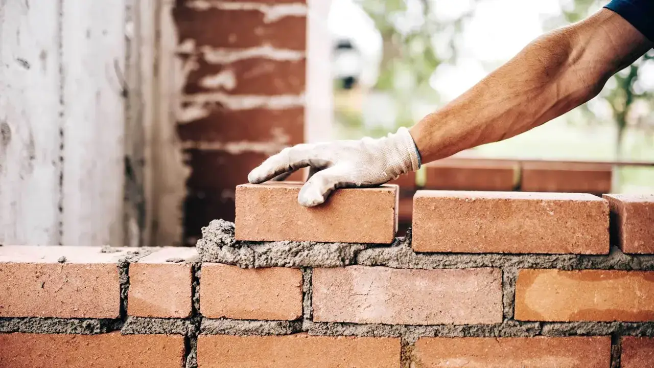 Ręka w rękawicy układa cegłę, kontynuując murowanie ścian działowych.