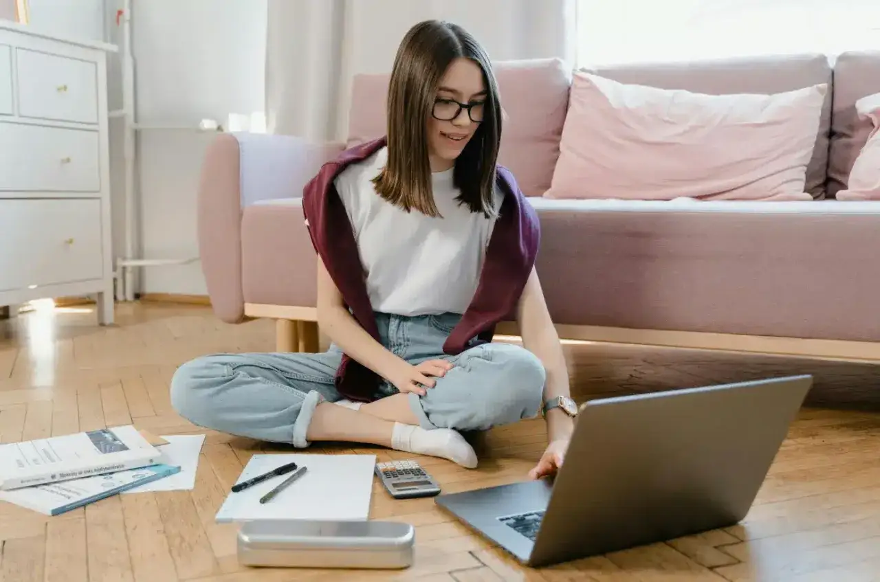 Dziewczyna uczy się matematyki online, siedząc na podłodze z laptopem i notatkami.