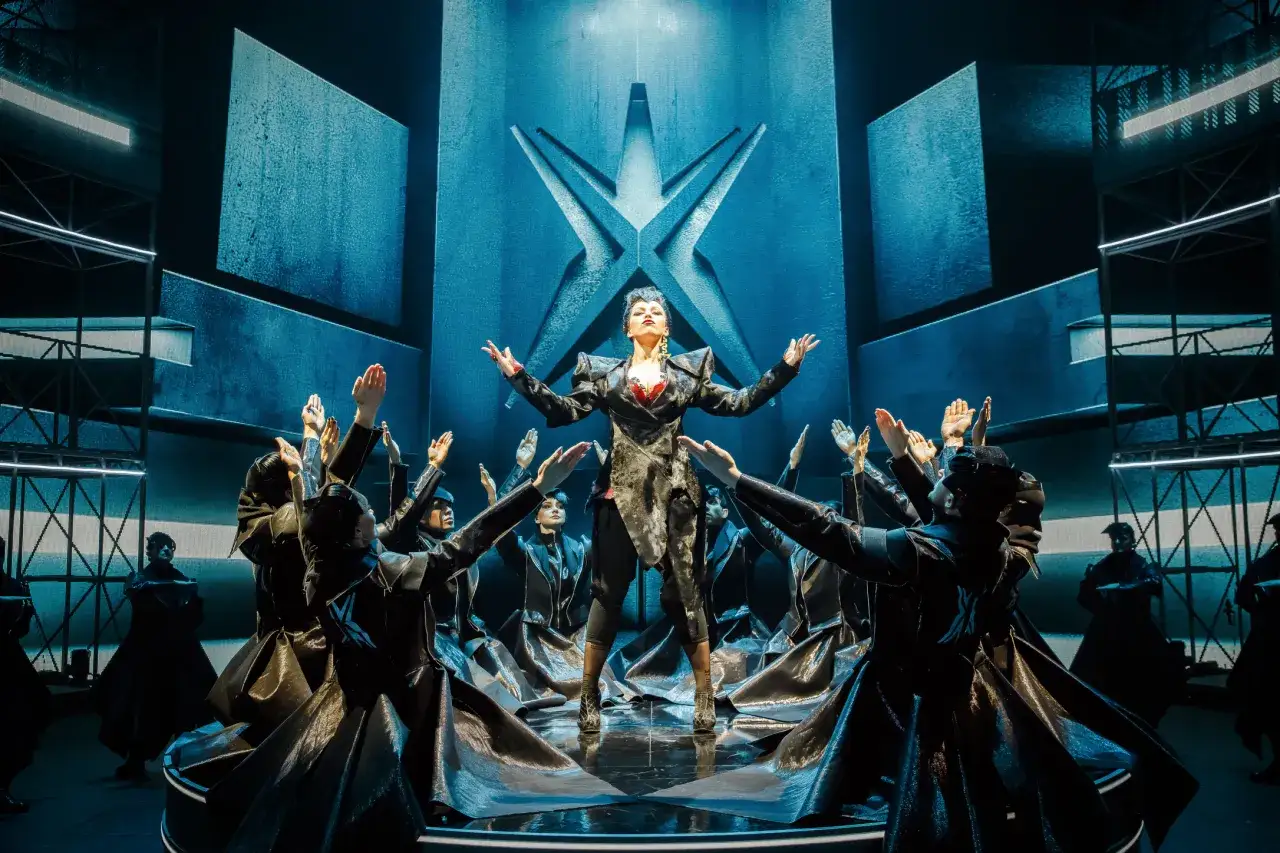 Wokalistka na scenie otoczona przez tancerzy w czarnych strojach. Spektakularna scenografia z niebieskimi światłami i gwiazdą na tle. To część repertuaru teatru Roma.