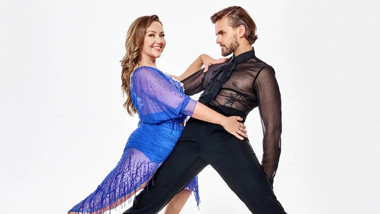 Para w zmysłowej pozie, kobieta w niebieskiej sukience z frędzlami, mężczyzna w prześwitującej koszuli. Ich ciała tworzą dynamiczną ramę w tańcu.