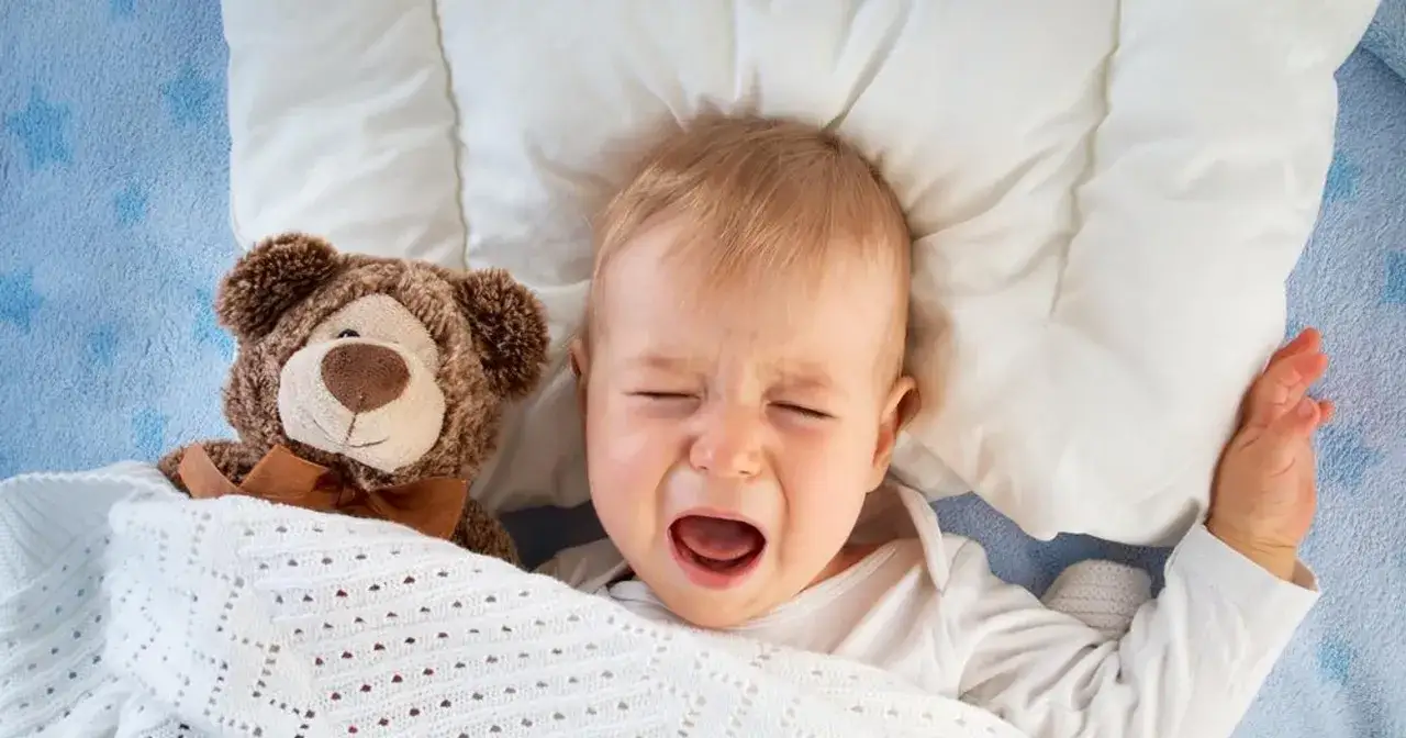 Płacz niemowlaka z suchym kaszlem. Maluch leży w łóżeczku z misiem, przykryty białym kocykiem.