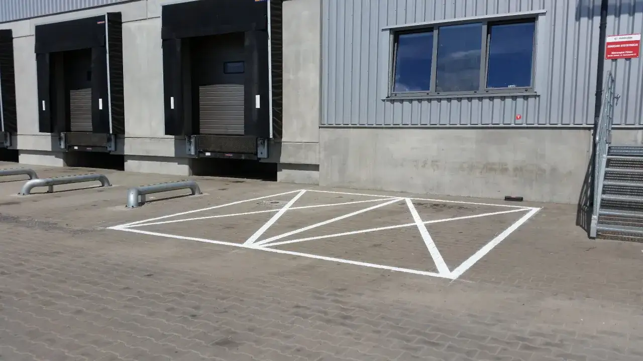 Parking z kopertą oznacza miejsce zarezerwowane dla osób niepełnosprawnych. Na zdjęciu widać takie oznaczenie na placu przed budynkiem magazynowym.