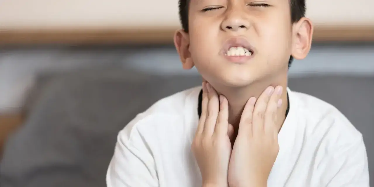Chłopiec z zamkniętymi oczami i grymasem na twarzy trzyma ręce na gardle, szukając odpowiedzi na pytanie: co na chrypę u dziecka.