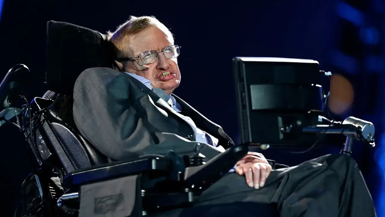 Stephen Hawking, wybitny naukowiec na wózku, w garniturze, z charakterystycznym uśmiechem i okularami, przemawia do publiczności.