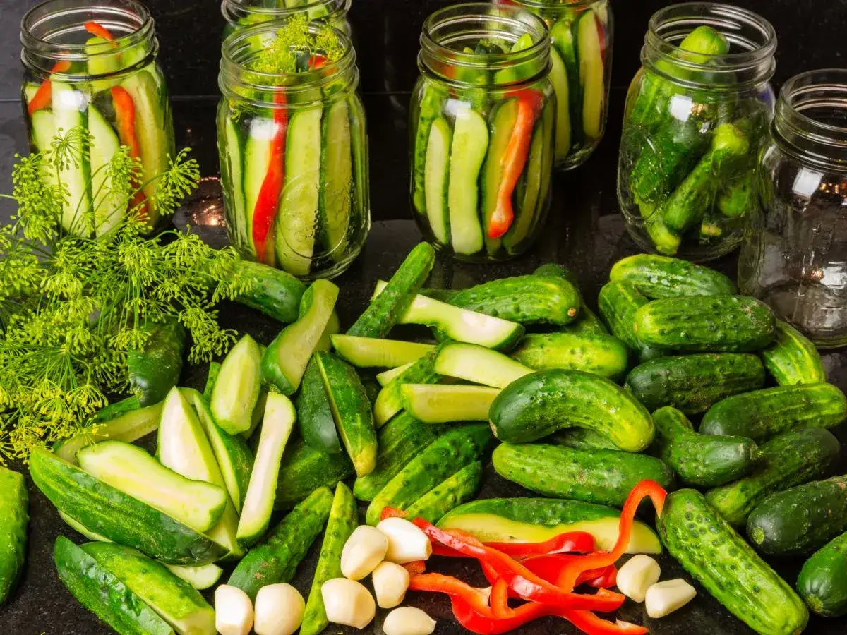 Słoiki z ogórkami, koperkiem i papryką, gotowe na pikle z ogórków. Na blacie leżą świeże ogórki, czosnek i kawałki papryki.