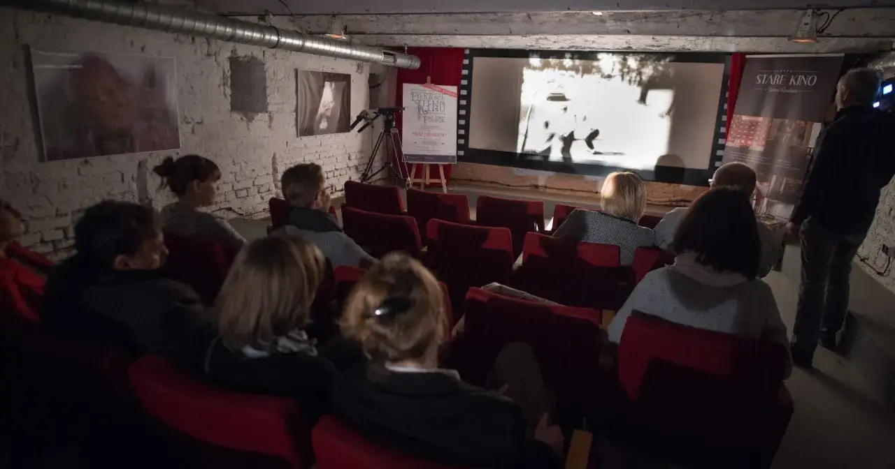 Widownia w kameralnym kinie ogląda film. Na ekranie widać scenę z epoki, a obok plakat informujący o premierze 