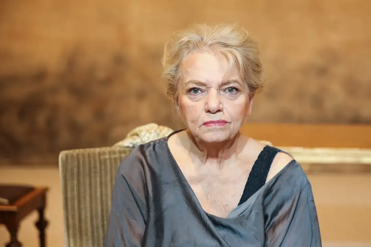 Anna Seniuk, obecnie, siedzi na krześle w szarej bluzce.