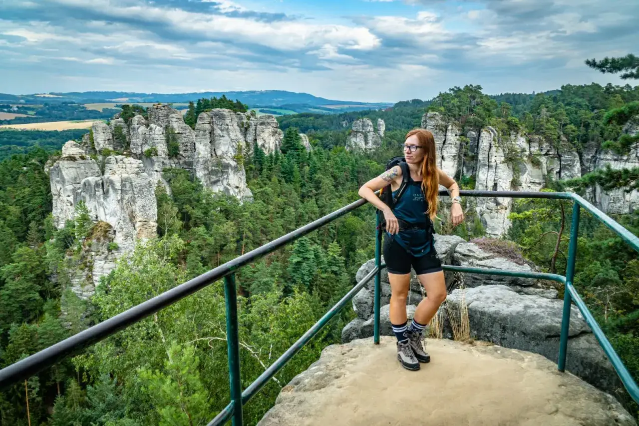 Kobieta w okularach i z plecakiem odpoczywa na punkcie widokowym, podziwiając imponującą, hrubą skalę otoczoną zielonym lasem.
