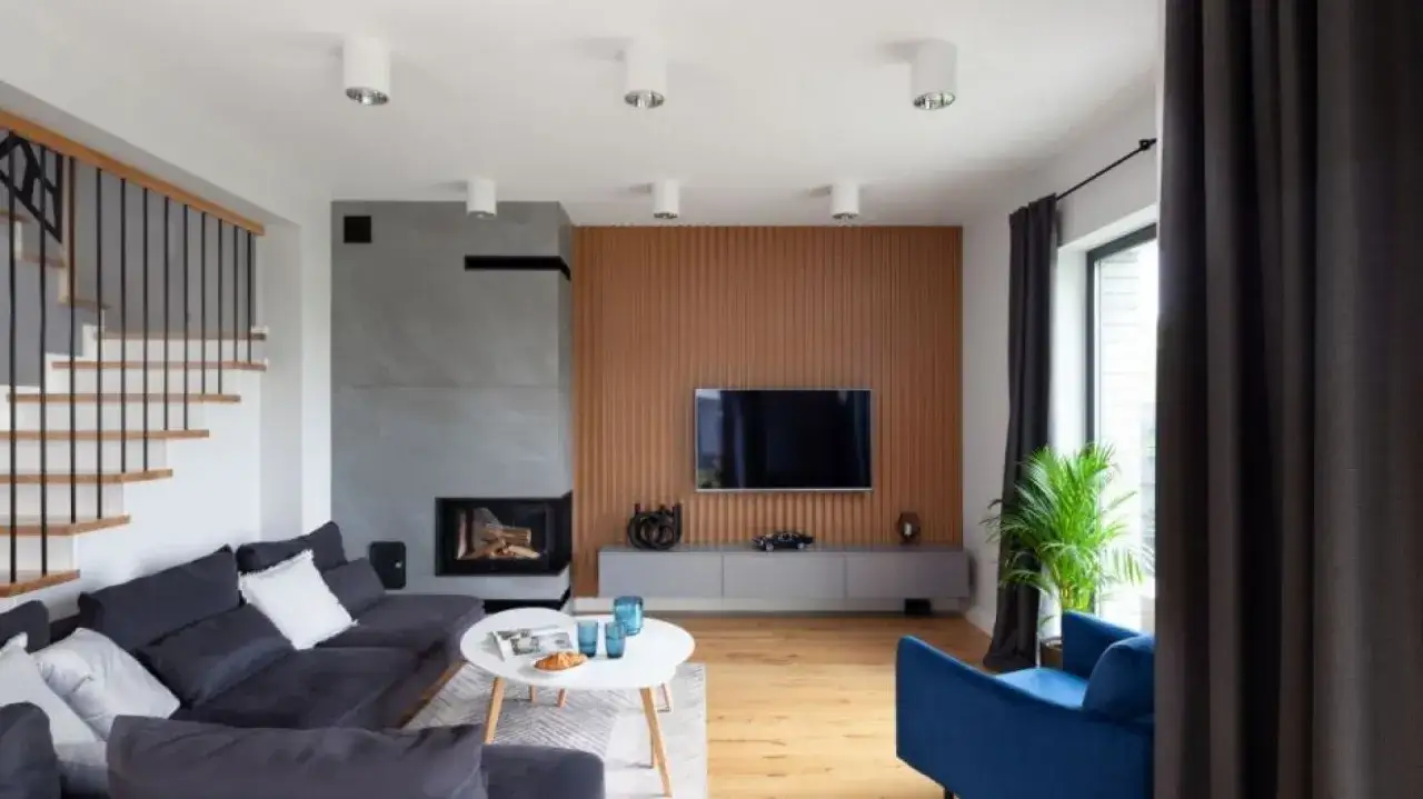 Nowoczesny salon z szarą sofą, białym stolikiem i palącym się kominkiem w salonie. Obok schody i telewizor na drewnianej ścianie.