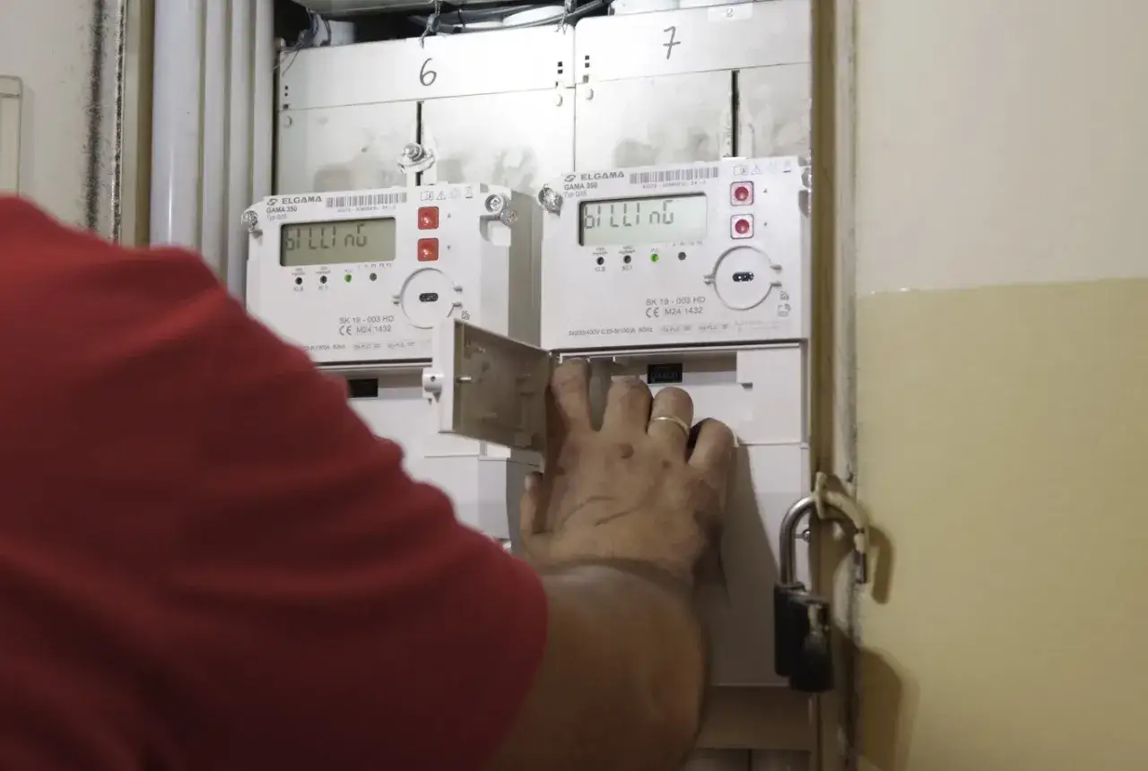 Osoba w czerwonej bluzce wykonuje odczyt licznika elektrycznego. Widoczne dwa liczniki z cyfrowymi wyświetlaczami.