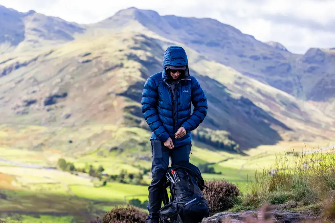 Mężczyzna w niebieskiej kurtce z kapturem, z plecakiem i kijkami trekkingowymi, na tle g&oacute;r. Taka kurtka to świetny przykład, co to jest **Primaloft** &ndash; lekka, ciepła i wodoodporna.