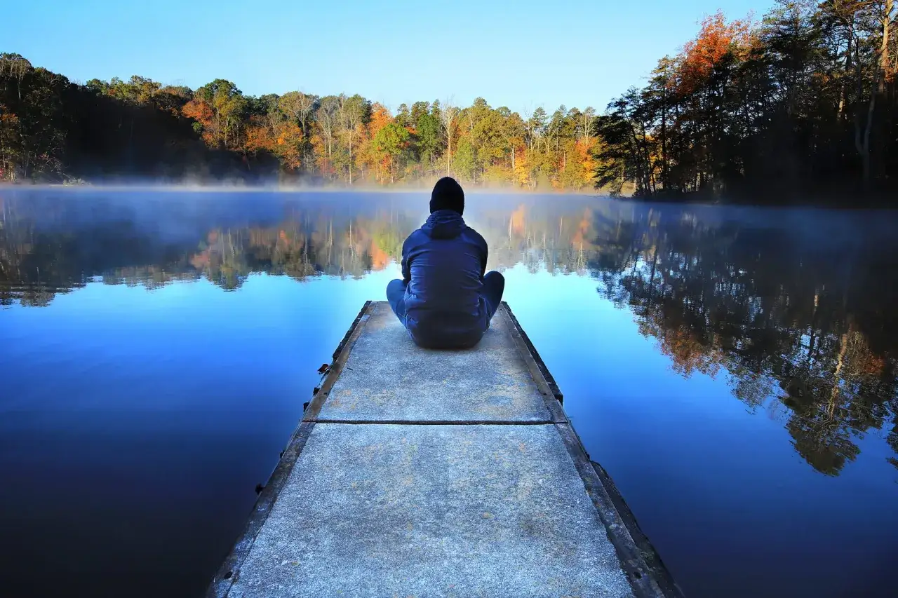 O poranku na pomoście siedzi osoba, kt&oacute;ra chce być sama. Mgła unosi się nad spokojną wodą, odbijając jesienne drzewa.