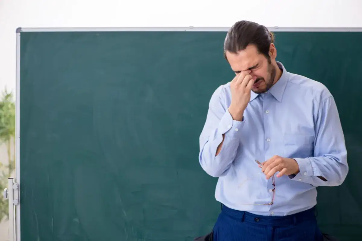 Zmęczony nauczyciel z bólem głowy, trzymający okulary, stoi przed tablicą. Czyżby dodatek za wychowawstwo był zbyt obciążający?