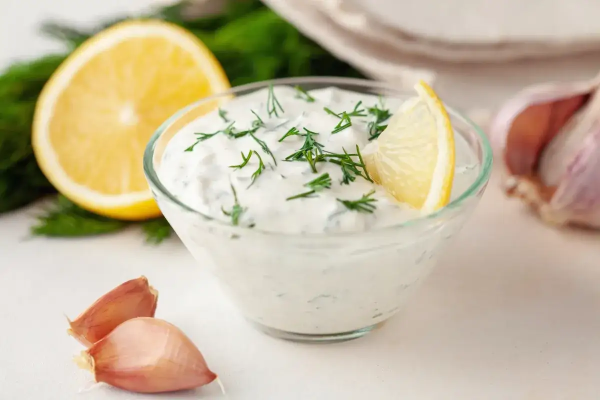 Pyszny sos koperkowy w szklanej miseczce, udekorowany plasterkiem cytryny i koperkiem. Obok czosnek i połówka cytryny.