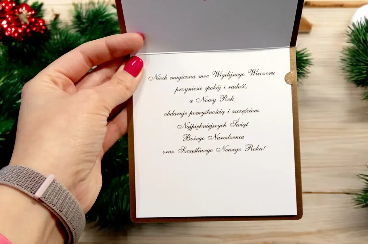 Oficjalne życzenia bożonarodzeniowe na eleganckiej kartce. Dłoń z r&oacute;żowym lakierem do paznokci trzyma otwartą kartkę z tekstem.