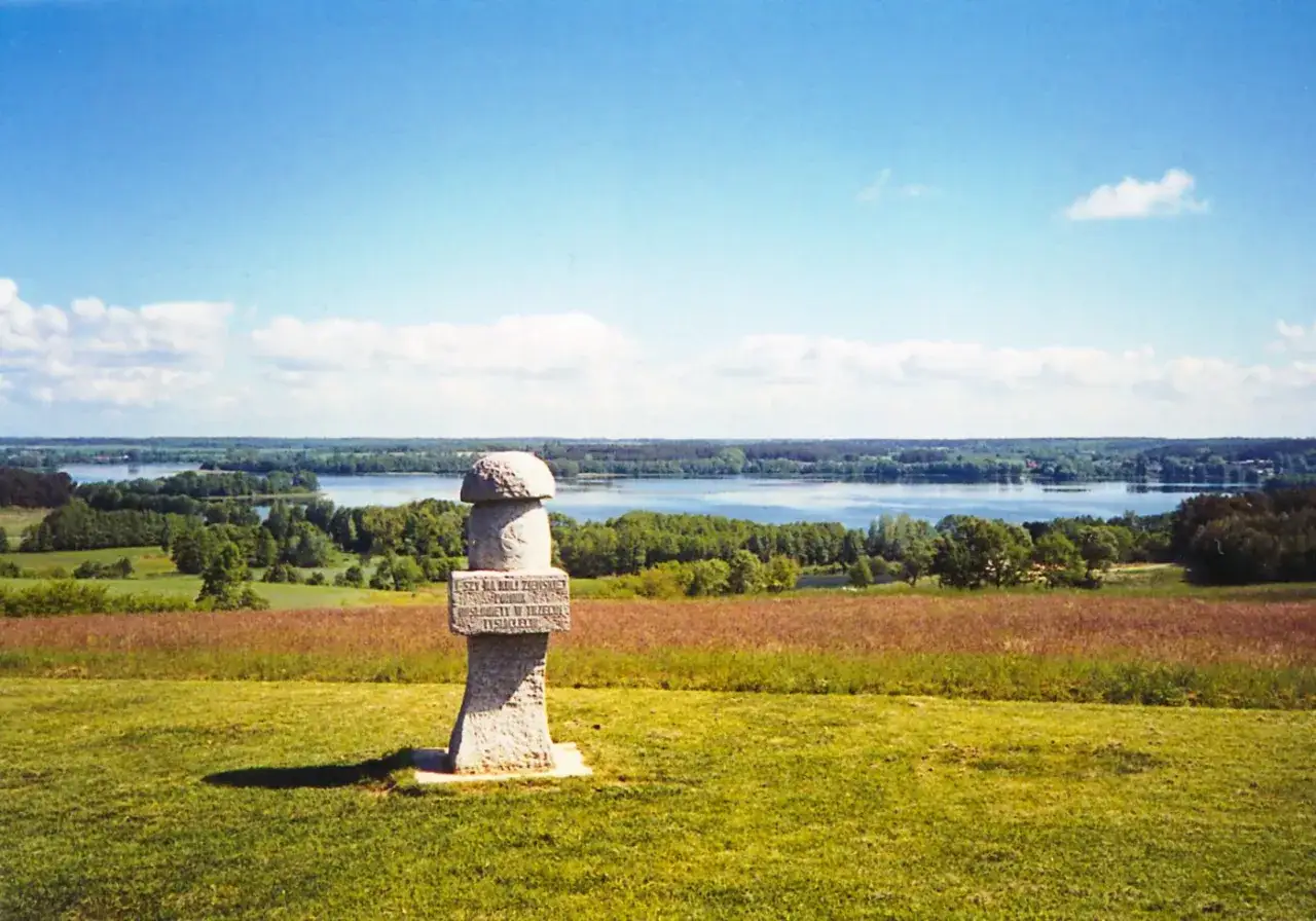 Kamienny obelisk na wzgórzu zapowiada piękno szlaku stu jezior. W oddali rozciąga się spokojna tafla wody otoczona zielenią.