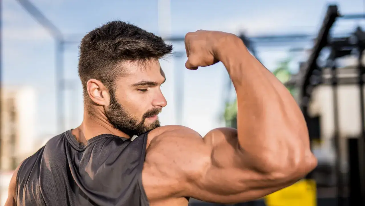 Mężczyzna z brodą napina potężny biceps, demonstrując efekty ćwiczeń na mięsień ramienny.