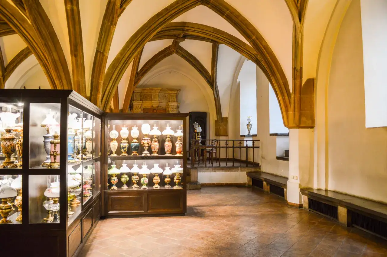 Wnętrze muzeum mieszczaństwa we Wrocławiu z zabytkowymi lampami naftowymi w gablotach i gotyckimi sklepieniami.