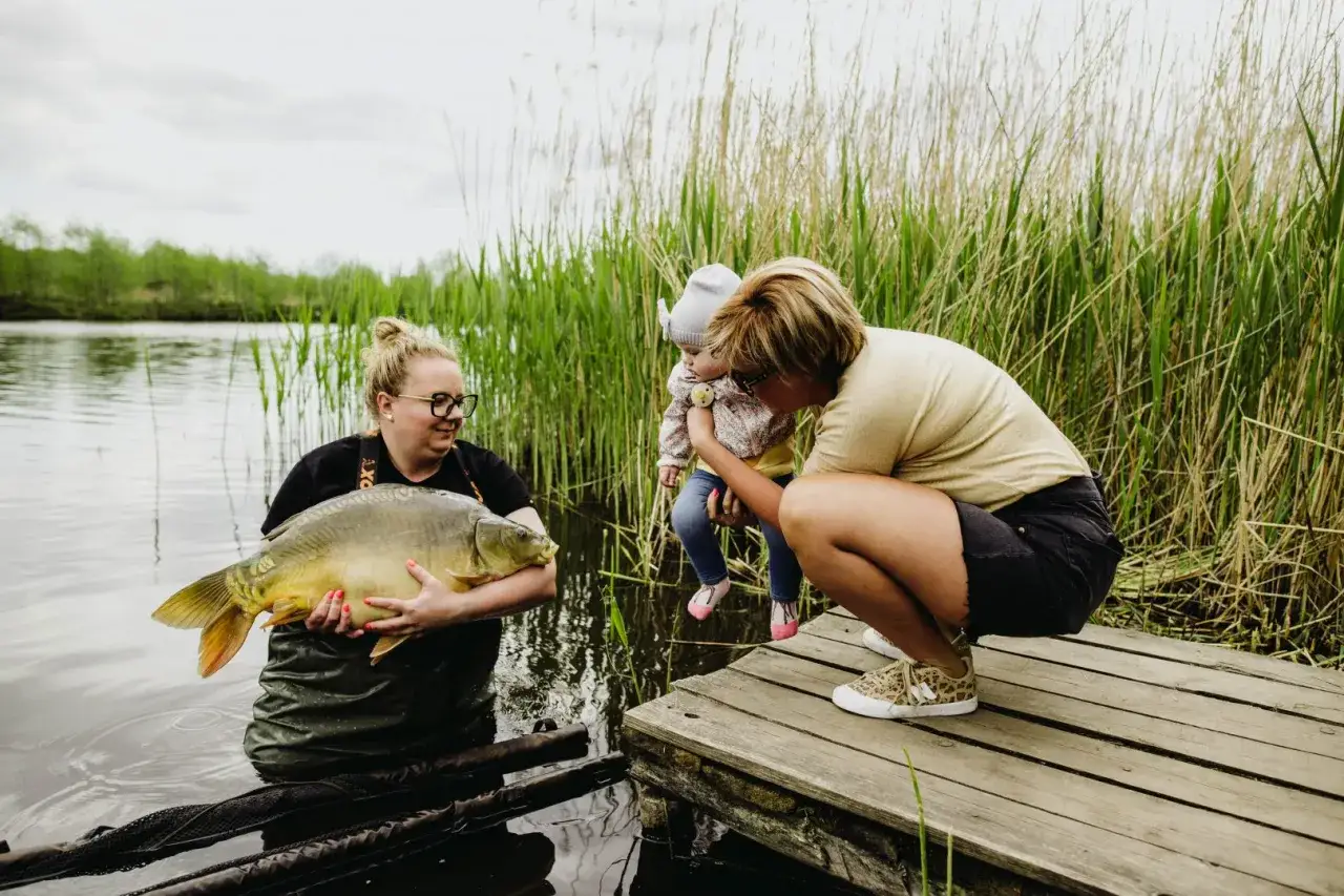 Rodzina na łowisku borowy staw. Kobieta trzyma dużą rybę, druga kobieta pokazuje ją dziecku.