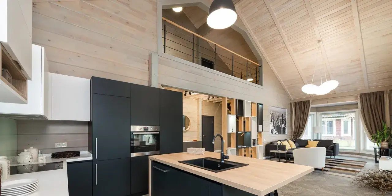 Nowoczesne lampy na poddasze skosy oświetlają przestronne wnętrze z drewnianymi belkami i aneksem kuchennym.