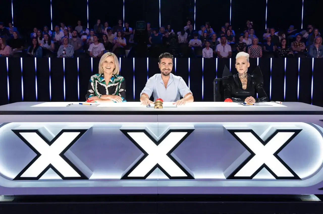 Trzyosobowe jury, w tym jedna kobieta z tatuażami, siedzi za stołem z dużymi literami 
