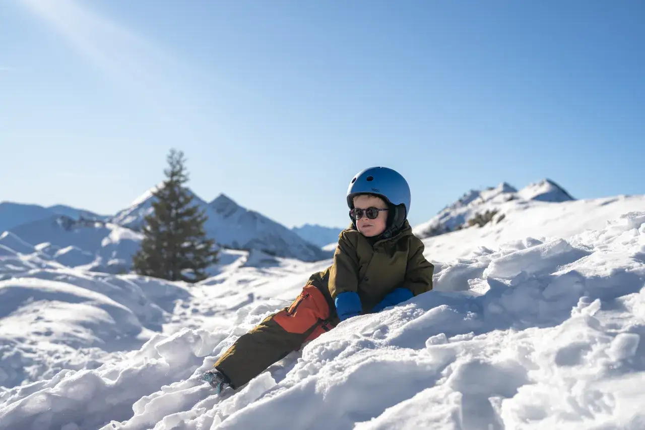 Mały chłopiec w niebieskim kasku i goglach odpoczywa na śniegu. W tle góry i choinka. Idealne miejsce na pierwsze narty dla 3 latka.